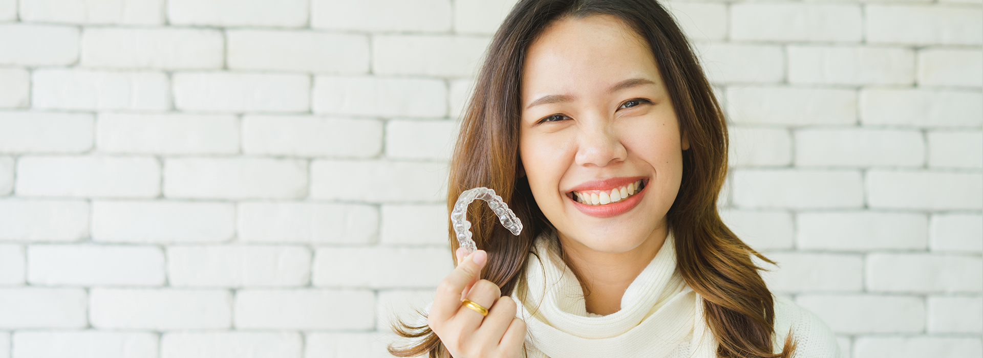 A woman wearing a white top and smiling, holding a ring in her hand against a brick wall.