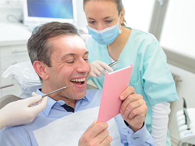 A man in a dental chair, holding a pink card and smiling at it, while a dentist with a blue mask looks on.