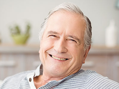 The image shows a smiling older man with gray hair, wearing a blue shirt and sitting in a chair.